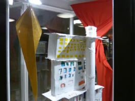 Ausstellung in einer Glasvitrine, Kunstwerke aus Stoff, bemaltes Hochhaus aus Pappe.