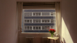 Filmstill von einem Raum mit Tisch und Blumenvase.