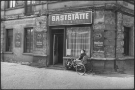 Eine Fotografie von einem Mann in einem Dreirad für Erwachsene vor einer Gaststätte.