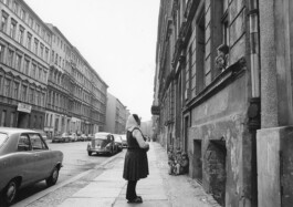 Eine Fotografie zeigt eine Frau auf dem Bürgersteig, die mit Kindern spricht, die zum Fenster rausschauen.