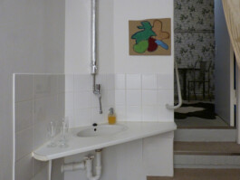 Das Waschbecken im Ausstellungsraum, daneben hängt eine Stickerei auf Pappe.