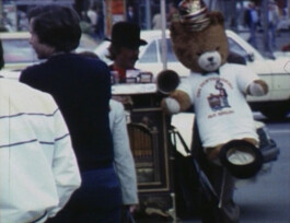 Filmstill einer Straßenszene in Berlin mit Passant*innen und einem Drehorgelspieler mit Teddybär