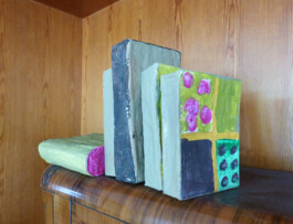 Bunt bemalte Bücher aus Pappmache stehen in einem Holzregal.