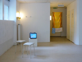 Ausstellungsraum mit Stühlen, Fernseher und einem Kunstwerk das aus Handtüchern hergestellt wurde.